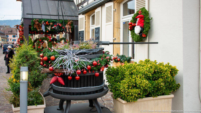 Large planter filled with evergreen branches, red ornaments, silver twigs, and a bow beside a decorated building façade.