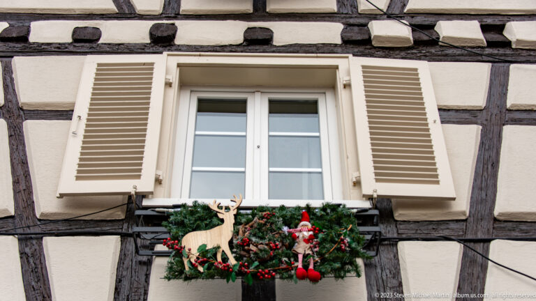 White-shuttered window in a half-timbered wall with a Christmas box holding evergreen, red ornaments, Santa, and a wooden reindeer.