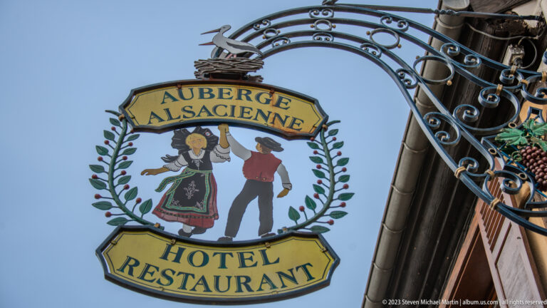 Ornate hanging sign reading “Auberge Alsacienne – Hotel Restaurant,” with a painted Alsatian couple and grapevine border.