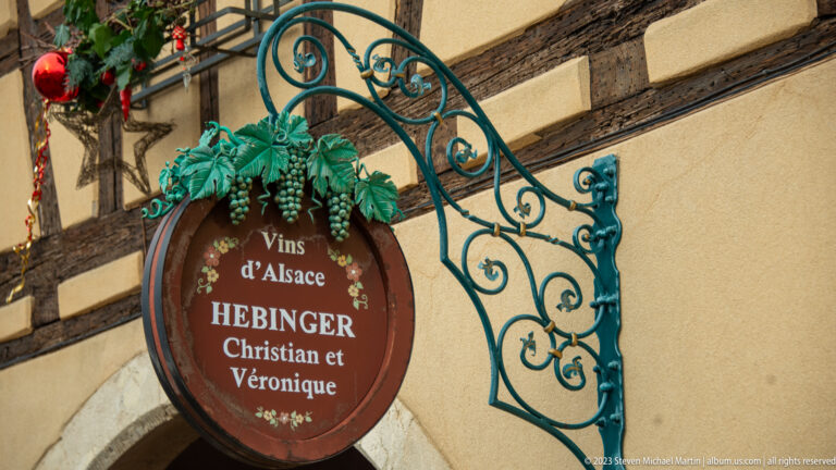 Round “Vins d’Alsace” sign for Hebinger Christian et Véronique hanging from an ornate green metal bracket with grape decoration.