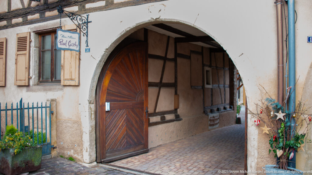 Large plastered arch with open wooden door leading into a narrow timber-framed, cobbled passage decorated for Christmas.