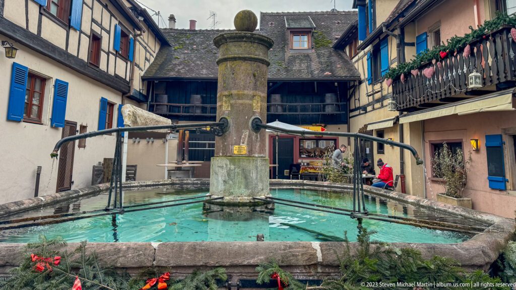 Rectangular stone fountain basin with a central column and curved rails, enclosed by traditional houses and balconies.
