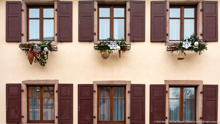 Pale residential façade with two rows of brown-shuttered windows and Christmas greenery decorating the upper flower boxes.