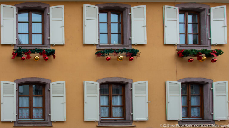 Ochre house façade with pale green shutters and upper window boxes filled with evergreen, red, and gold Christmas ornaments.