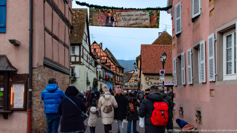 Crowd in winter coats walking along a narrow historic street in Eguisheim beneath a festive overhead banner.