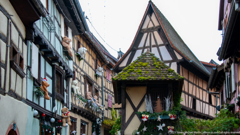 Narrow Eguisheim lane framed by steep-roofed half-timbered houses, façade Christmas decorations, and a small roofed structure in the foreground.