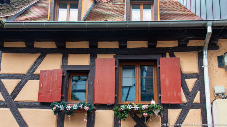 Half-timbered beige façade with two windows, weathered red shutters, and small Christmas-decorated flower boxes.