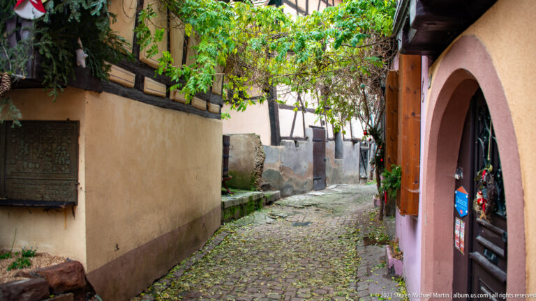 Narrow cobbled passage curving between old plastered walls and timber framing, with an arched doorway on the right.