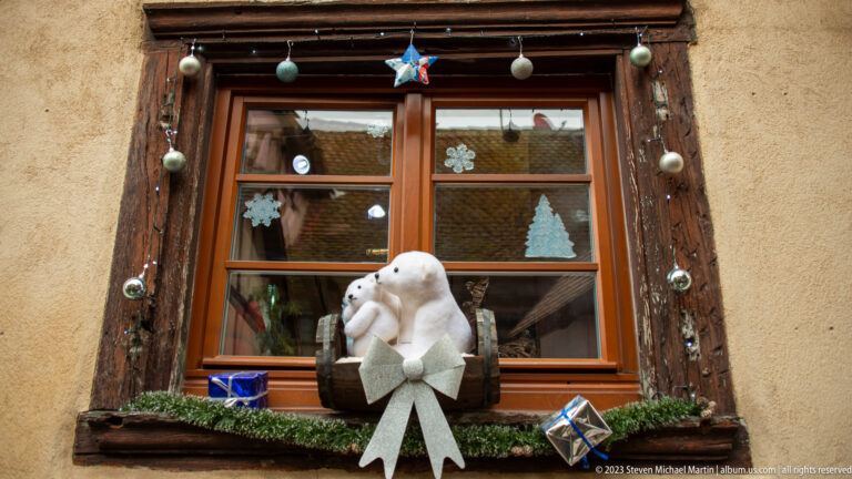 White bear decoration with a large bow sitting in a window box beneath a wood-framed window with hanging Christmas ornaments.