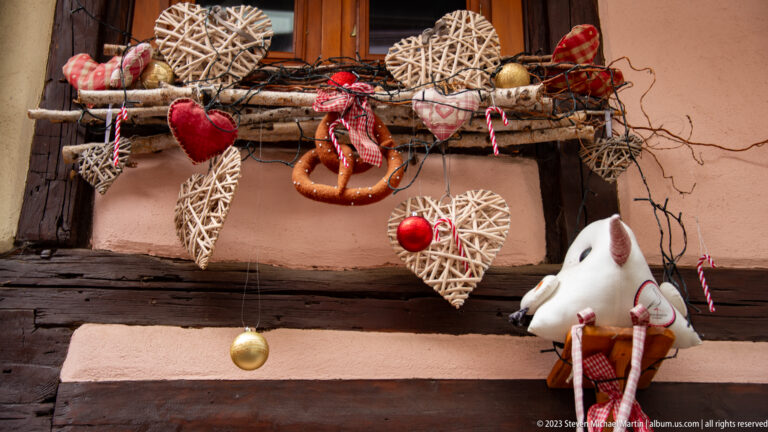 Rustic Christmas display with wicker hearts, red ornaments, pinecones, a pretzel, and a plush figure hanging from a timber beam.