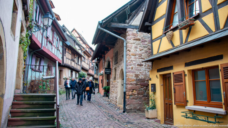 Cobblestone lane with pedestrians between colorful half-timbered houses, exposed stone, and projecting upper stories in Eguisheim.