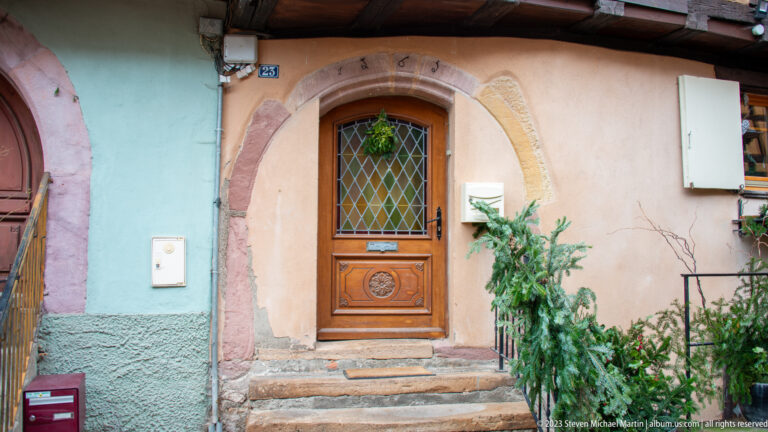 Carved wooden door with diamond-paned upper glazing set in a rounded stone arch, reached by shallow steps.