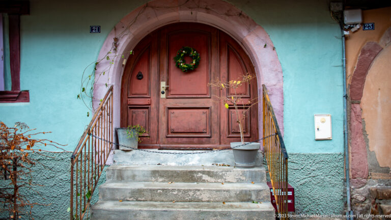 Reddish double wooden door with a Christmas wreath in a pink stone arch, reached by steps with simple iron railings.