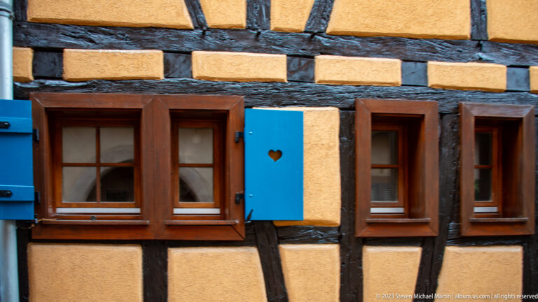 Yellow half-timbered wall with small wood windows and bright blue shutters, including one with a heart-shaped cutout.