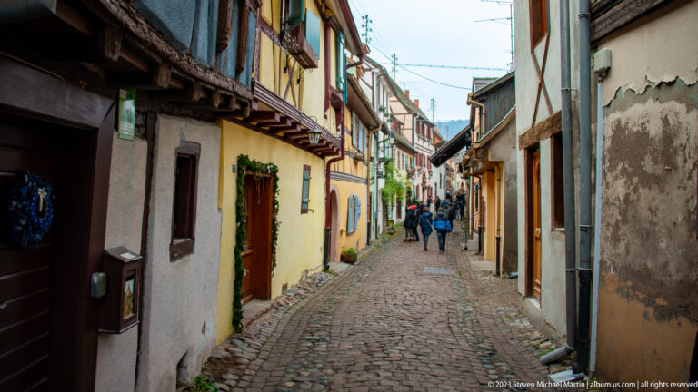 Long narrow cobblestone lane lined with historic plastered houses and a few pedestrians in the distance.