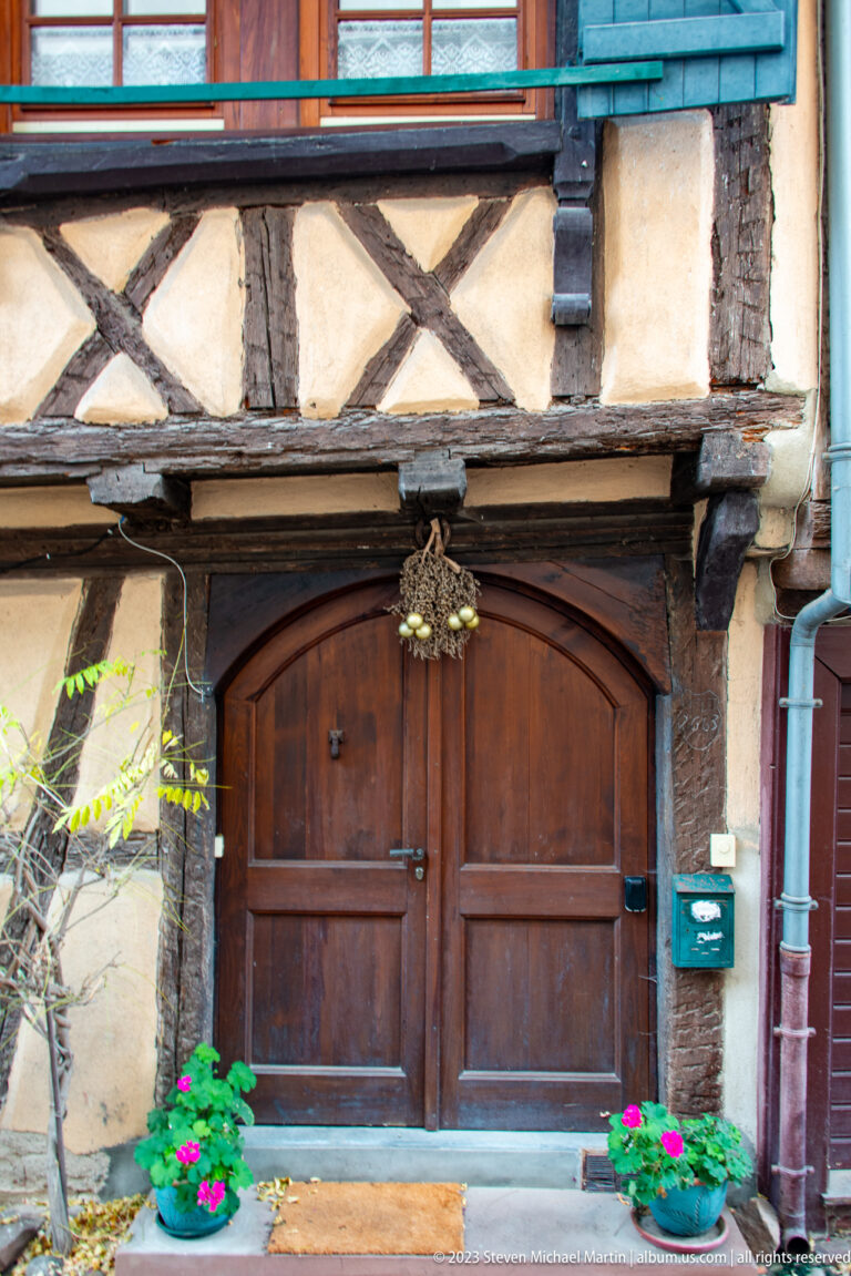 Wide dark wooden double door beneath exposed half-timber framing, with potted red flowers flanking the entrance.