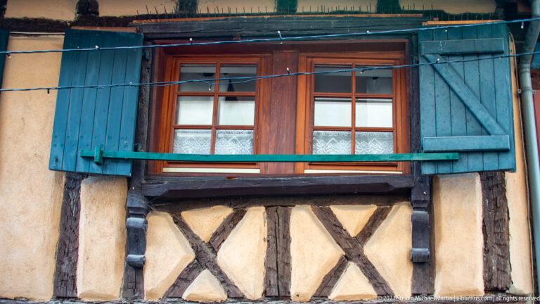 Wood-framed double window with weathered blue shutters above exposed dark timbers and pale plaster infill.