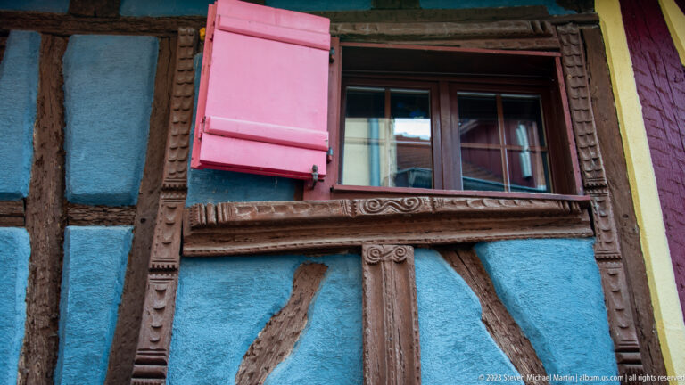 Window in a blue half-timbered wall with carved wooden surround and one bright pink shutter opened to the side.