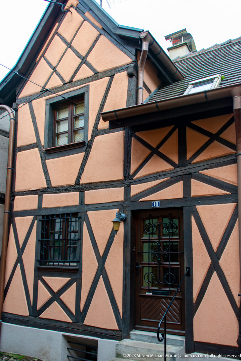 Compact peach-colored half-timbered house with dark diagonal bracing, small windows, sloping roofs, and a street-level entrance.