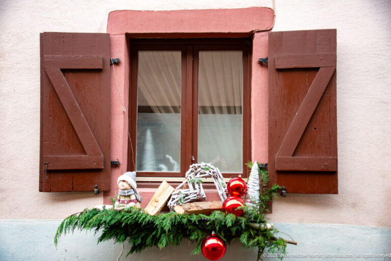 Brown-shuttered window with evergreen, a red bauble, small holiday figurines, and rustic decorations arranged on the sill.