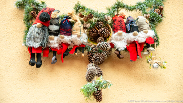 Row of plush gnome-like Christmas figures hanging with evergreen branches and pinecones against a pale yellow wall.