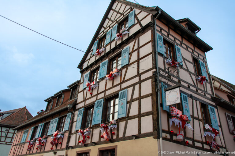 Steep-gabled half-timbered corner house with turquoise shutters and red-and-white Christmas decorations at the windows.