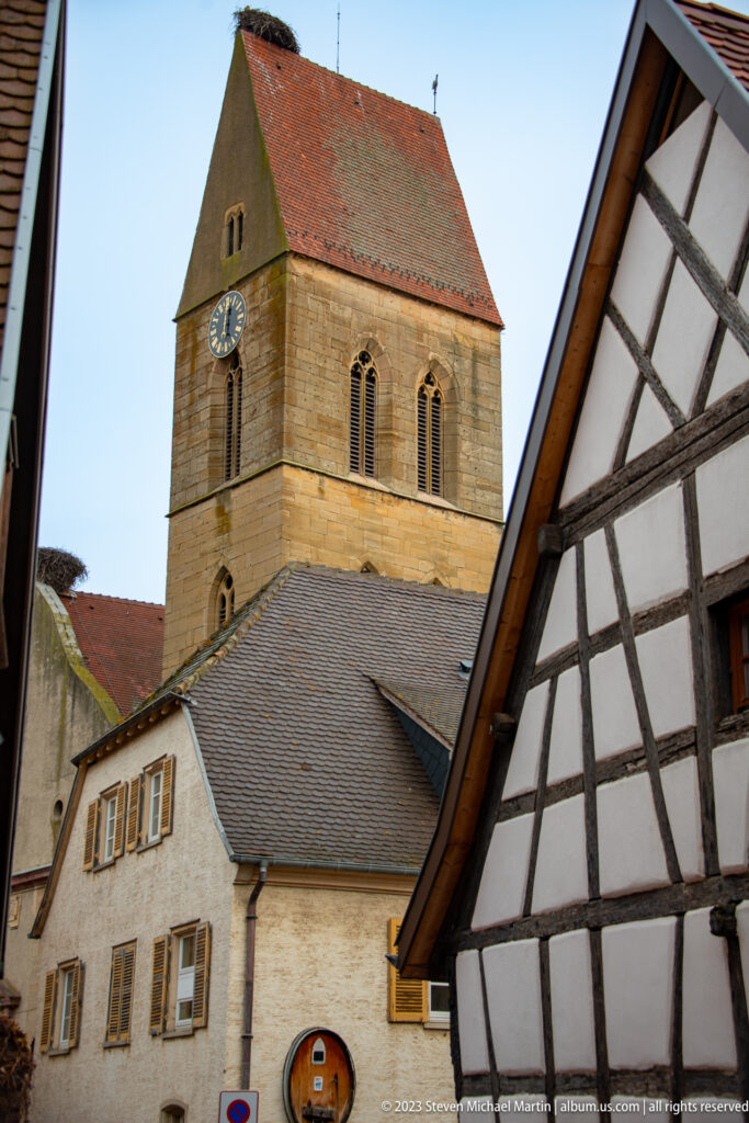 Square stone church tower with clock and arched openings rising behind tiled roofs and half-timbered houses.