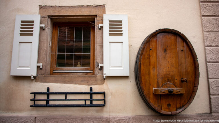 Oval wooden door with iron hardware beside a divided-glass window with white shutters in a pale plaster wall.