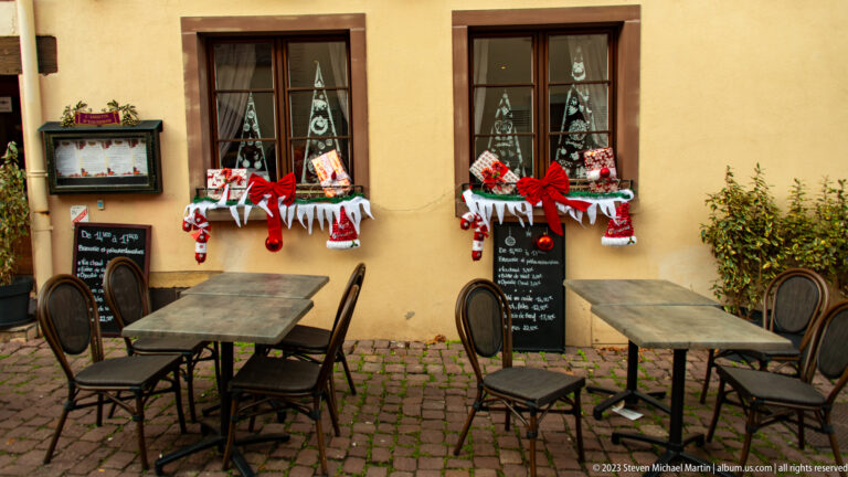 Outdoor café tables and chairs beneath two windows decorated with red bows, stockings, ornaments, and Christmas figures.