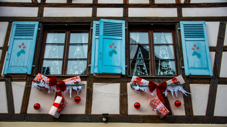 Two windows with turquoise floral shutters, red bows, stockings, gift ornaments, and white Christmas trees behind the glass.