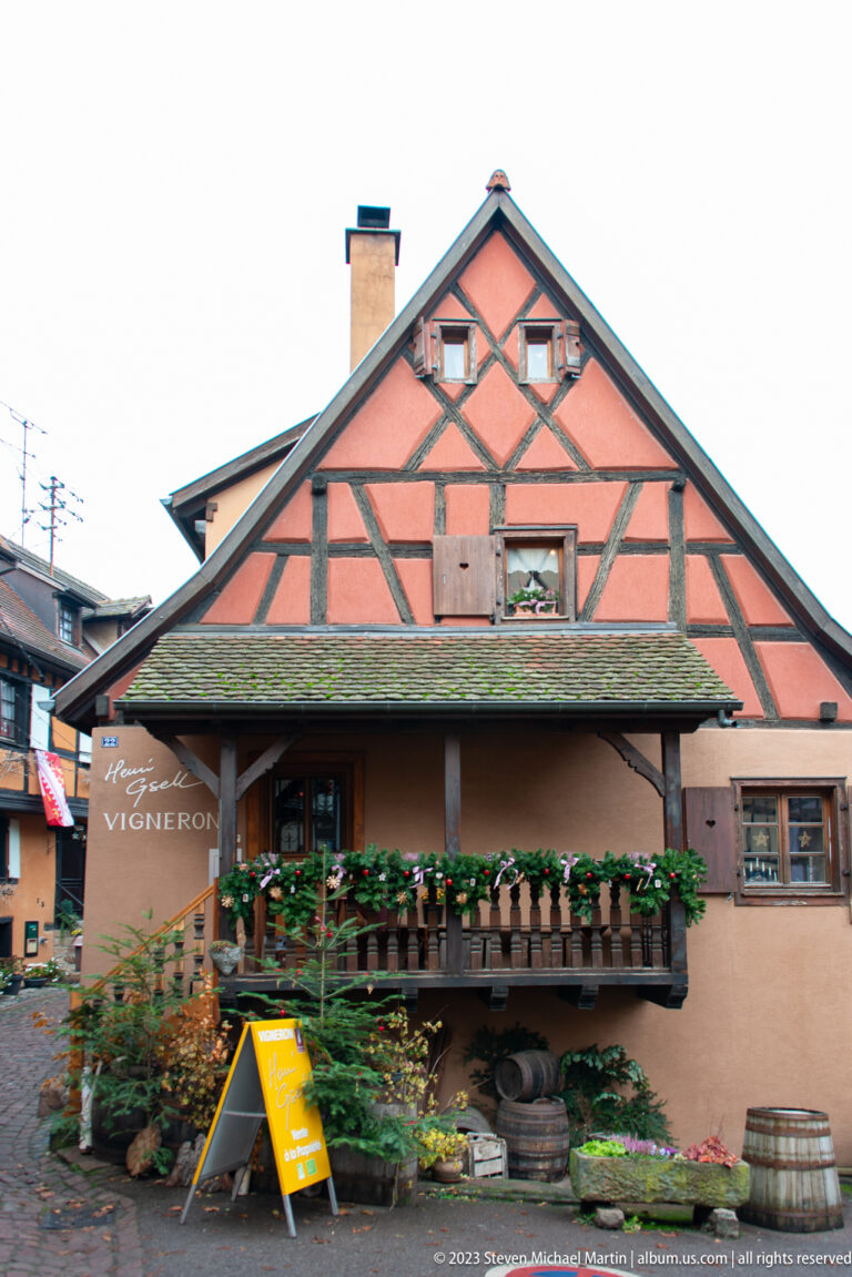 Salmon-colored half-timbered winegrower building with long balcony, flower boxes, barrels, and the word “VIGNERON” on the façade.