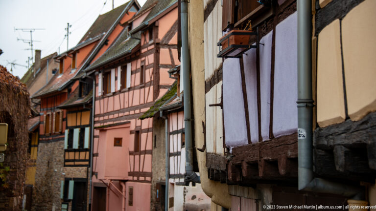 Narrow lane lined with pink, ochre, and pale half-timbered houses with projecting upper stories and tiled roofs.