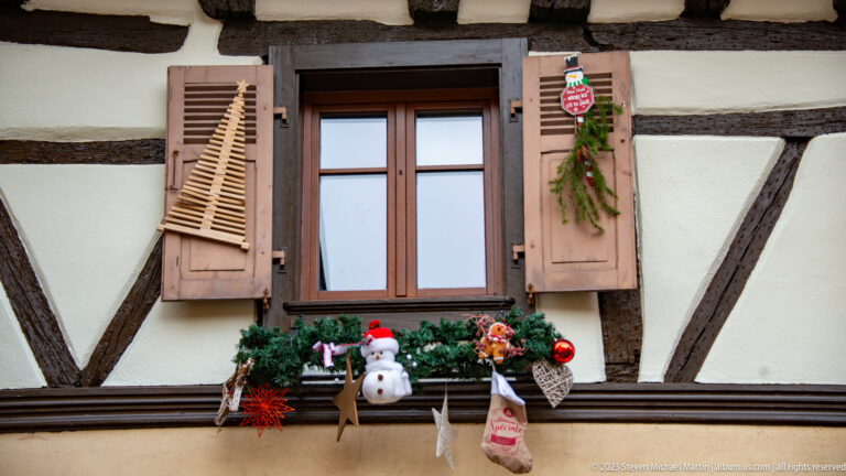 Brown-shuttered window with evergreen box, snowman, red ornaments, hanging figures, and a small shield decoration.