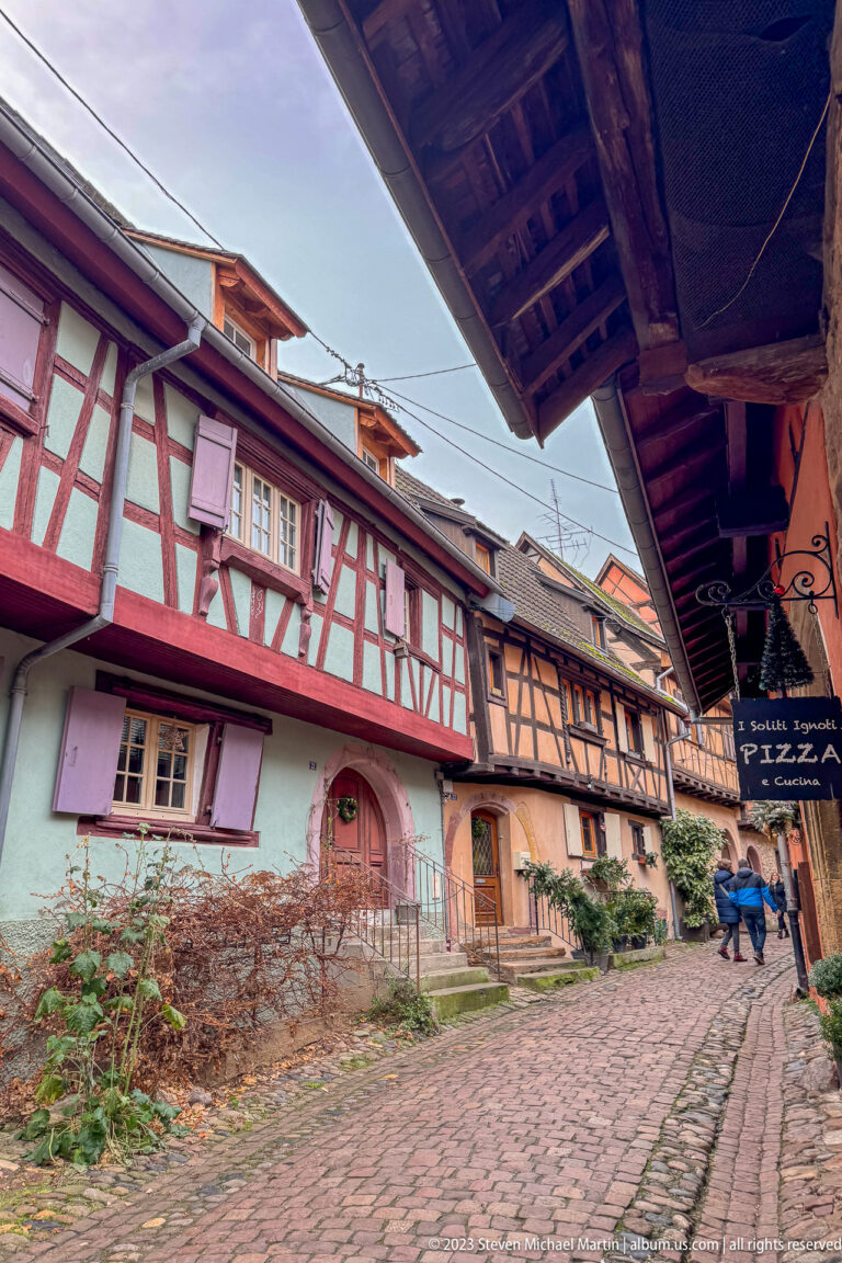 Curving cobbled lane bordered by pastel half-timbered houses, deep eaves, and a projecting restaurant sign.