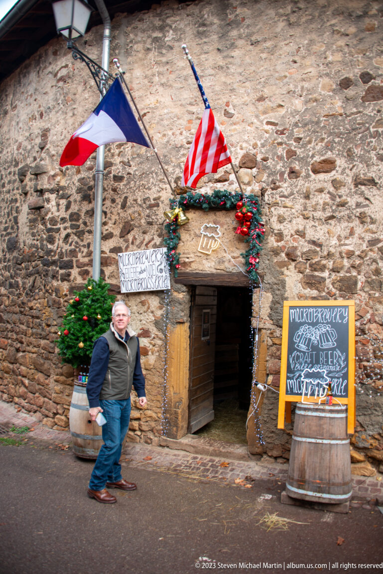Low stone microbrewery entrance decorated with French and American flags, evergreen, and chalkboard beer signage.