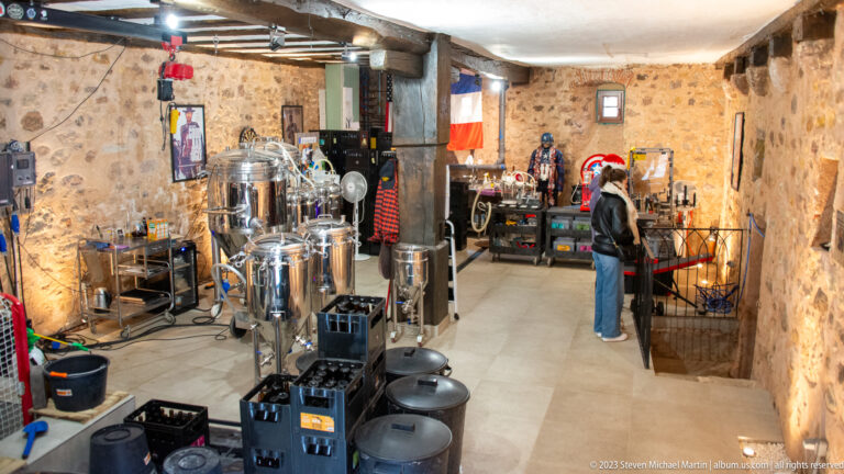 Stone-walled microbrewery interior with stainless-steel brewing vessels, hoses, kegs, work equipment, and a visitor.