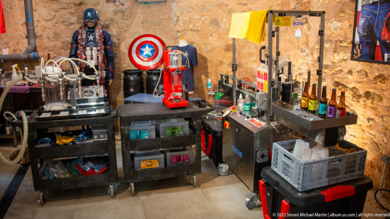 Microbrewery work station with bottles, processing equipment, crates, rolling carts, and a red machine against a stone wall.