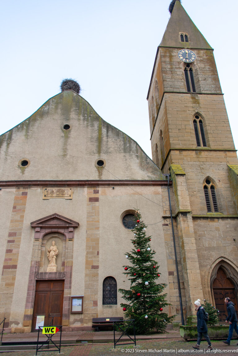 Church façade with sculptural niche and Christmas tree beside a square stone clock tower in Eguisheim.