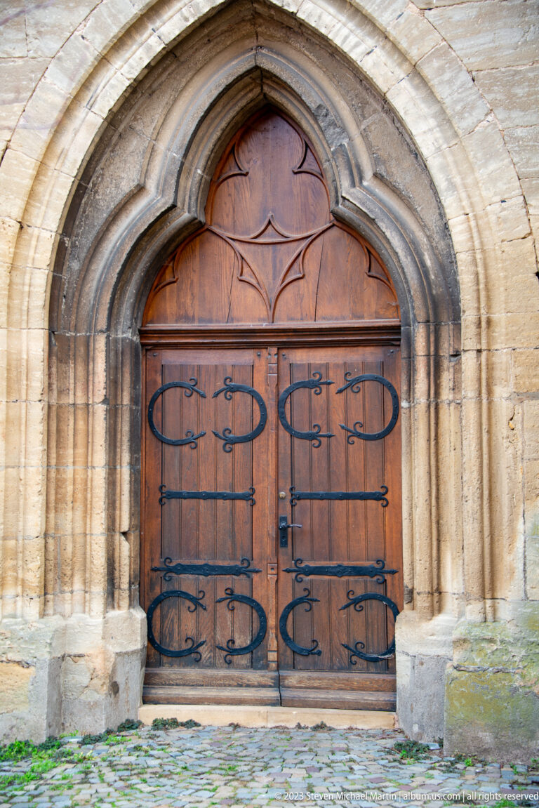 Pointed molded stone church portal with wooden double doors, decorative iron strap hinges, and cobbled threshold.