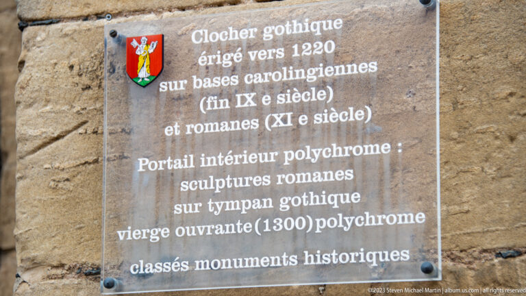 French information plaque describing a Gothic bell tower, earlier foundations, Romanesque sculpture, and a polychrome church portal.