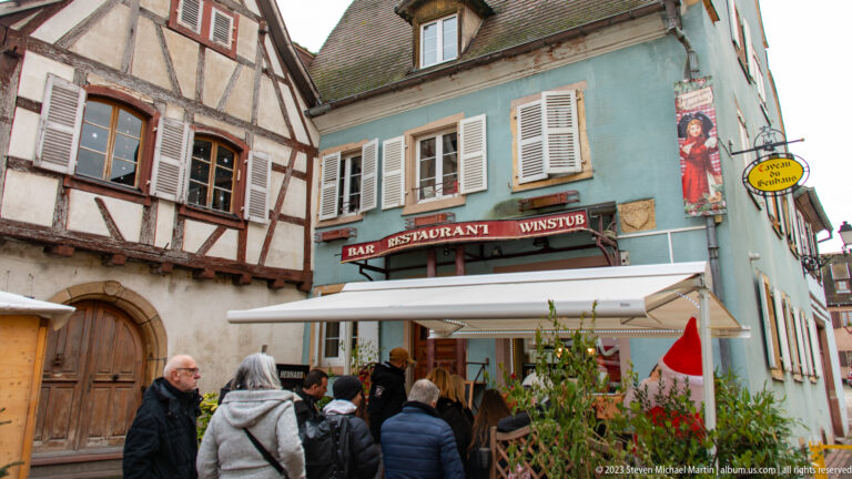 Visitors outside a blue-green corner building marked “BAR RESTAURANT WINSTUB,” beside a half-timbered house with arched doorway.