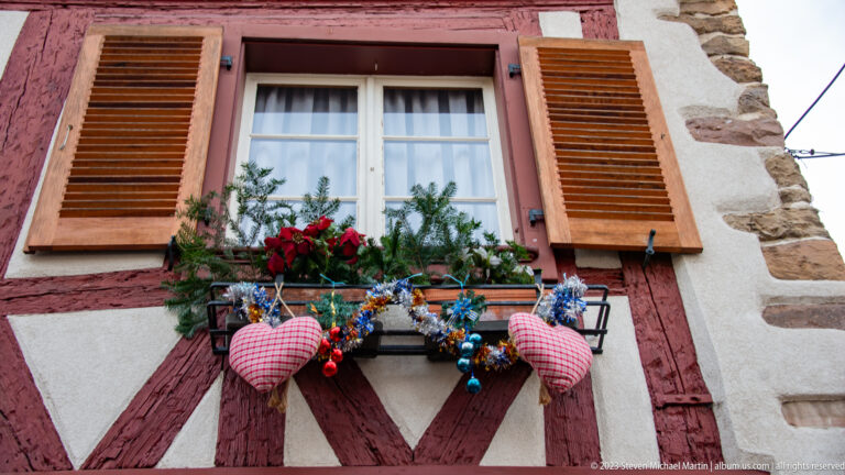 Window in red half-timber framing with brown shutters, evergreen, red flowers, blue ornaments, and two gingham heart decorations.