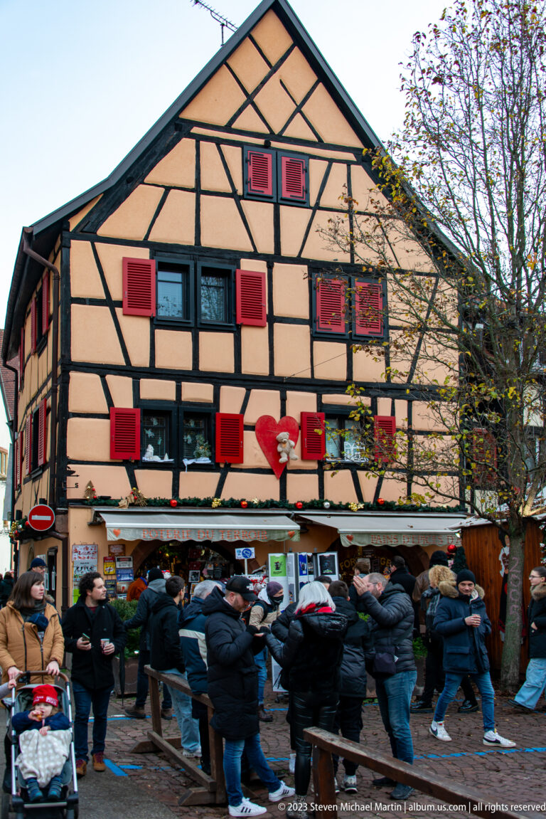 Winter crowd gathered at market counters beneath a peach half-timbered building with bright red shutters.
