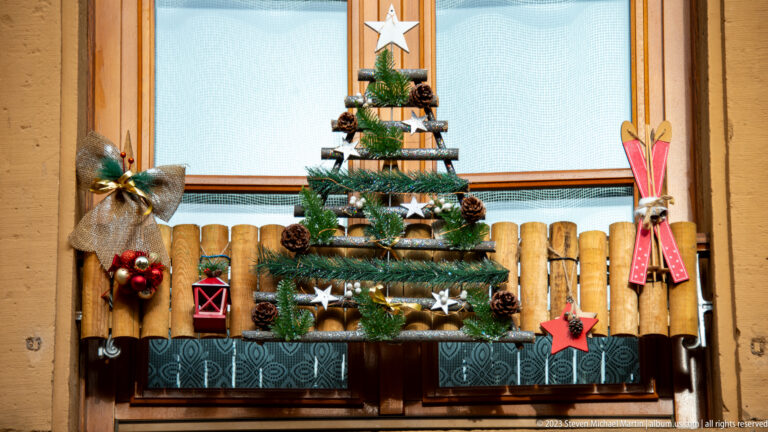 Wood-slat Christmas tree decorated with evergreen, pinecones, white stars, miniature red skis, and burlap ornaments on a window ledge.
