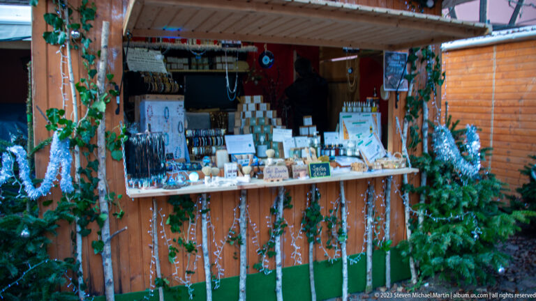Wooden Christmas market booth decorated with evergreen and silver tinsel, displaying jewelry, small crafts, and boxed goods.