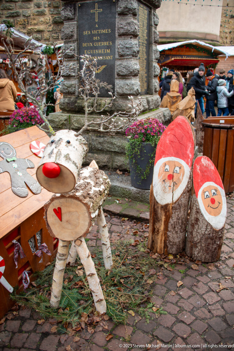 Handmade birch-log reindeer beside upright logs painted with Santa faces on cobbled ground at the Christmas market.