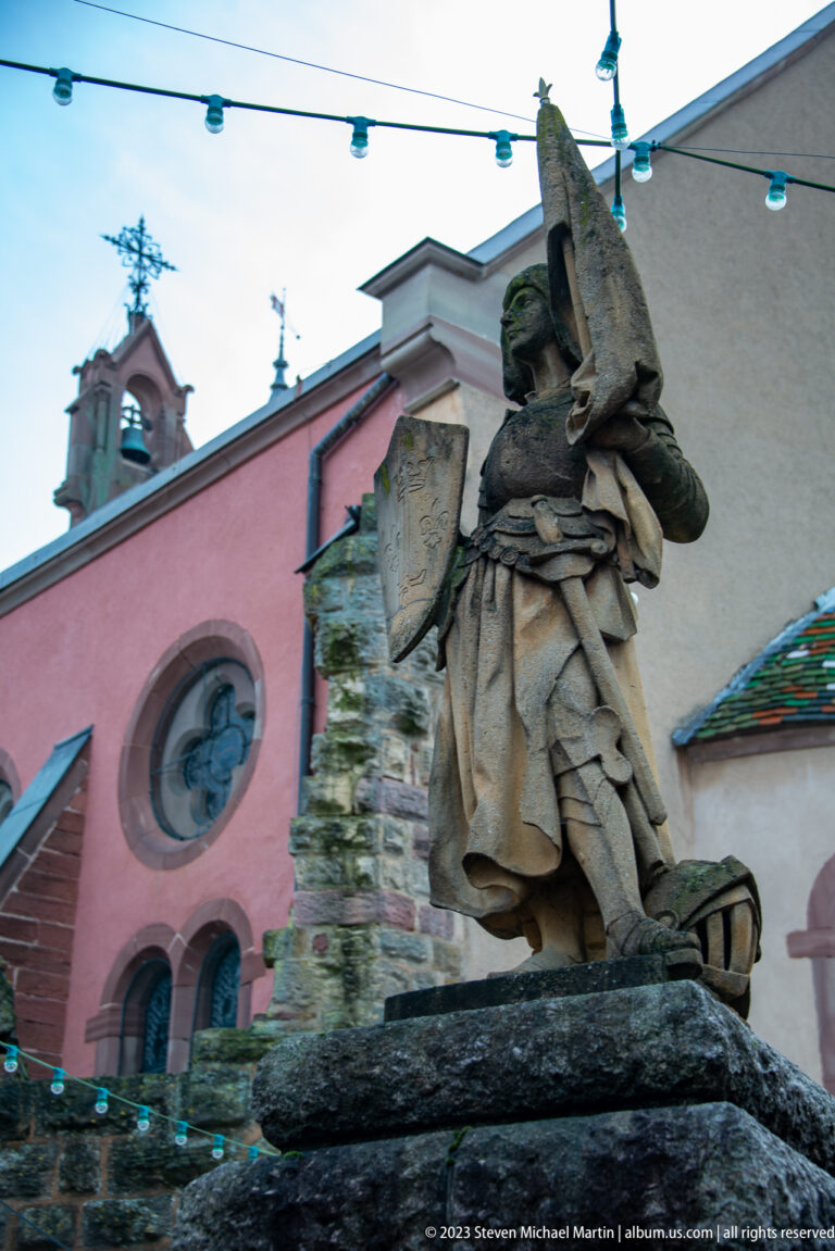 Weathered stone statue of a robed figure on a sculpted pedestal, viewed from below against a pink church façade.