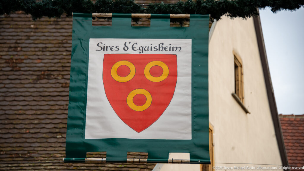 Green-bordered banner reading “Sires d’Eguisheim” above a red shield bearing three gold ring-like roundels.