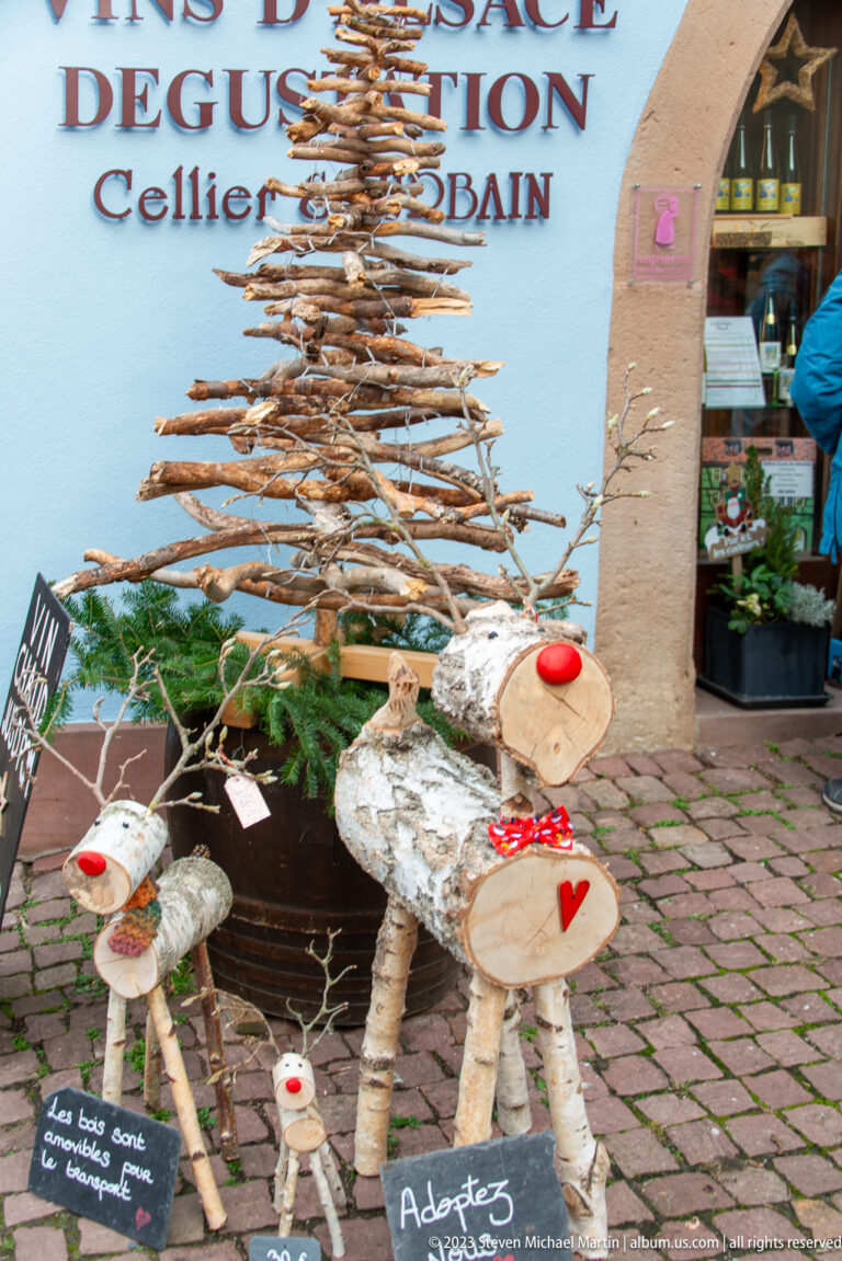 Birch-log reindeer with red noses and branch antlers standing beneath a tall twig Christmas tree on a cobbled street.