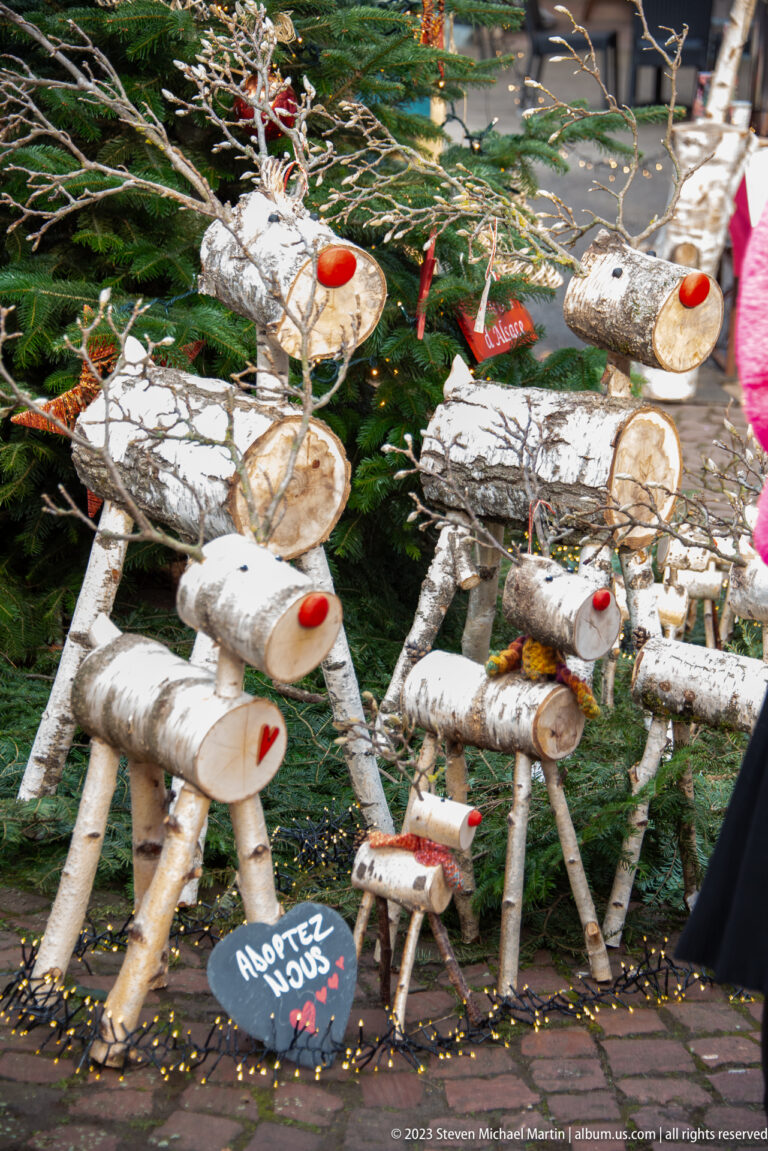 Group of handmade birch-log reindeer with twig antlers and red noses beside a small sign reading “Adoptez nous.”